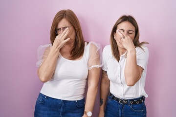 Hispanic mother and daughter wearing casual white t shirt over pink background smelling something stinky and disgusting, intolerable smell, holding breath with fingers on nose. bad smell - obrazy, fototapety, plakaty