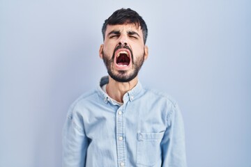 Young hispanic man with beard standing over blue background angry and mad screaming frustrated and furious, shouting with anger. rage and aggressive concept.