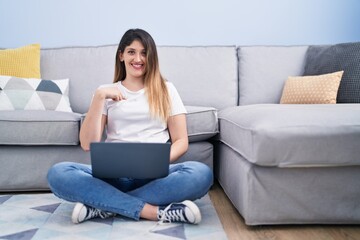 Fototapeta premium Young brunette woman sitting on the floor at home using laptop pointing finger to one self smiling happy and proud