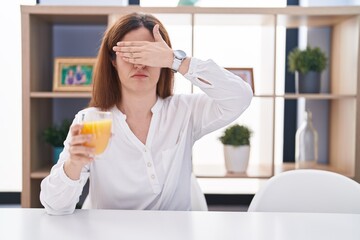 Brunette woman drinking glass of orange juice covering eyes with hand, looking serious and sad. sightless, hiding and rejection concept