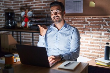 Hispanic man with beard working at the office at night smiling with happy face looking and pointing to the side with thumb up.