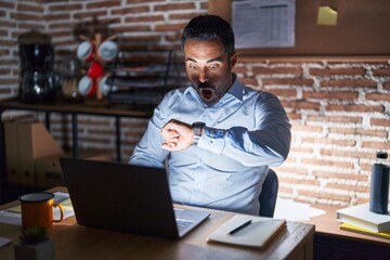 Hispanic man with beard working at the office at night looking at the watch time worried, afraid of getting late