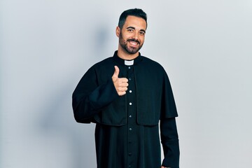 Handsome hispanic man with beard wearing catholic priest robe doing happy thumbs up gesture with hand. approving expression looking at the camera showing success.