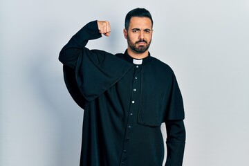 Handsome hispanic man with beard wearing catholic priest robe strong person showing arm muscle,...
