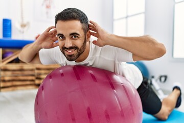 Young hispanic man patient smiling confident having rehab session using fit ball at clinic