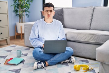 Non binary person studying using computer laptop sitting on the floor puffing cheeks with funny face. mouth inflated with air, crazy expression.
