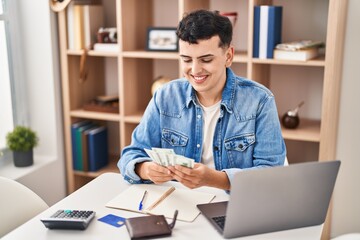 Young non binary man sitting on table counting dollars at home