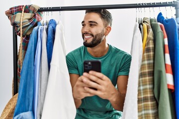 Young arab man customer using smartphone appearing through clothes rack at clothing store