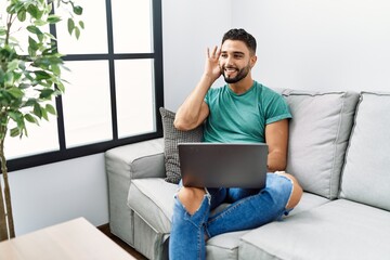 Young handsome man with beard using computer laptop sitting on the sofa at home smiling with hand over ear listening an hearing to rumor or gossip. deafness concept.