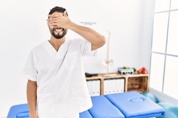 Fototapeta premium Young handsome man with beard working at pain recovery clinic smiling and laughing with hand on face covering eyes for surprise. blind concept.