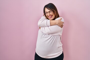 Pregnant woman standing over pink background hugging oneself happy and positive, smiling confident. self love and self care