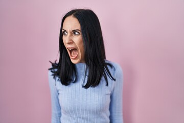 Hispanic woman standing over pink background angry and mad screaming frustrated and furious,...