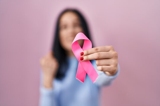 Hispanic woman holding pink cancer ribbon screaming proud, celebrating victory and success very excited with raised arms