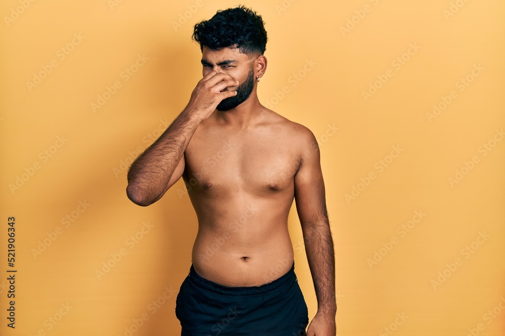 Poster arab man with beard wearing swimwear shirtless smelling something stinky and disgusting, intolerable