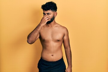 Fototapeta premium Arab man with beard wearing swimwear shirtless smelling something stinky and disgusting, intolerable smell, holding breath with fingers on nose. bad smell