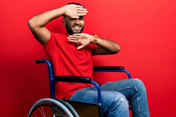 Arab man with beard sitting on wheelchair smiling cheerful playing peek a boo with hands showing...