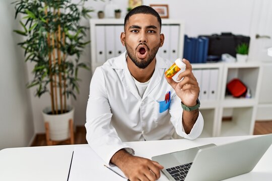 Young indian doctor holding prescription pills scared and amazed with open mouth for surprise, disbelief face