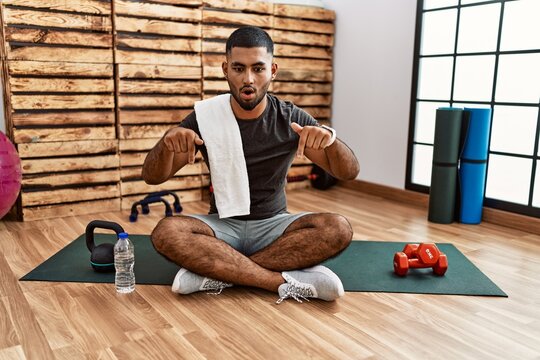 Young indian man sitting on training mat at the gym pointing down with fingers showing advertisement, surprised face and open mouth