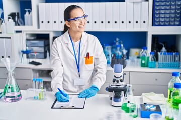 Fototapeta premium Young hispanic woman scientist writing on document working at laboratory