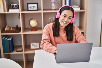 Young hispanic woman using laptop and headphones sitting on table at home