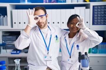 Interracial couple working at scientist laboratory smiling happy doing ok sign with hand on eye looking through fingers