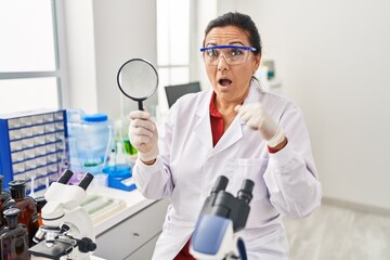 Middle age hispanic woman working at scientist laboratory holding magnifying glass scared and amazed with open mouth for surprise, disbelief face