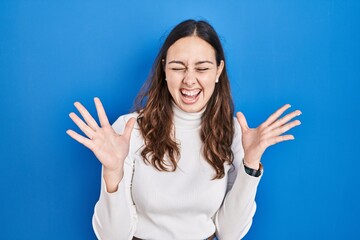 Fototapeta premium Young hispanic woman standing over blue background celebrating mad and crazy for success with arms raised and closed eyes screaming excited. winner concept