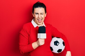 Young hispanic woman holding reporter microphone and soccer ball winking looking at the camera with...