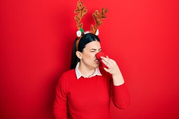 Young hispanic woman wearing deer christmas hat and red nose smelling something stinky and...