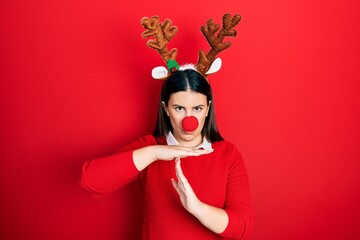 Young hispanic woman wearing deer christmas hat and red nose doing time out gesture with hands,...