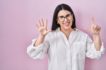 Young brunette woman standing over pink background showing and pointing up with fingers number seven while smiling confident and happy.