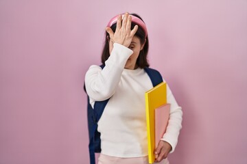 Woman with down syndrome wearing student backpack and holding books surprised with hand on head for...