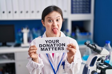 Woman with down syndrome working at scientist laboratory holding your donation matters banner sticking tongue out happy with funny expression.