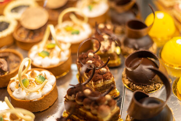 Rows of sweet pastries in a glass showcase at a patisserie. There are small round baked cakes, chocolate mousse, pastries, biscuits, berry tarts, and fruit snacks on display for sale. 