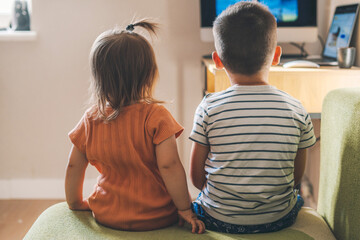 Back view of a brother with his baby sister watching television together in living room at home,...