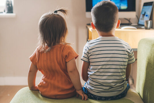 Back view of a brother with his baby sister watching television together in living room at home, sitting on the sofa. New generation and modern tech overuse