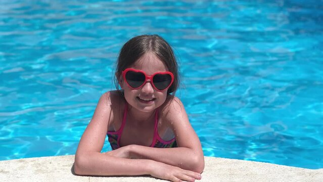Smiling cute little girl in heart shaped sunglasses in swimming pool on sunny day. Childhood, valentine's day and summer vacation concept. Happy kid having fun outdoors on holidays