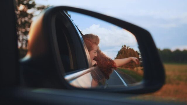 free woman travels by car catches wind with hand from car window. Girl with long hair is sitting in front seat of car, stretching her arm out window and catching glare of setting sun. Hand rays of sun