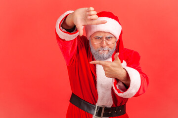 Fototapeta na wymiar Elderly man in santa claus costume gesturing picture frame with hands, looking through fingers and focusing on interesting moment, taking photo. Indoor studio shot isolated on red background.