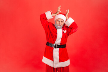 Fototapeta na wymiar I am deer! Elderly man with gray beard wearing santa claus costume showing deer antler horns over head, looking with comical humorous expression. Indoor studio shot isolated on red background.