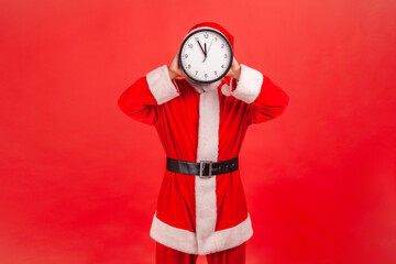 Portrait of unknown man in santa claus costume standing covering face with wall clock, wasting his...