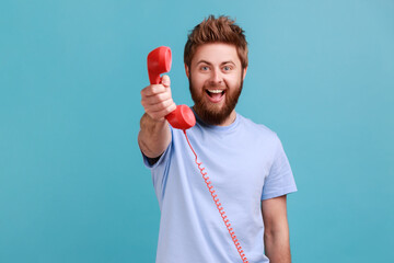 Portrait of smiling positive bearded man holding in hands red retro telephone, waiting for your call, looking at camera with excited expression. Indoor studio shot isolated on blue background.
