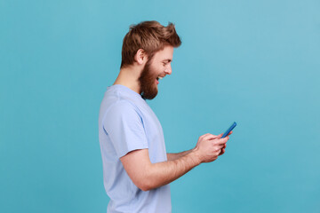 Fototapeta premium Side view of happy excited bearded man using online service on mobile phone and expressing pleasant emotions, texting messages for friends. Indoor studio shot isolated on blue background.