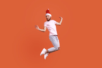 Fototapeta na wymiar Portrait of funny bearded man in santa hat jumping in the air and looking at camera with positive smile, spreading hands aside, wearing pink T-shirt. Indoor studio shot isolated on orange background.