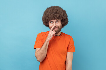 Portrait of childish man with Afro hairstyle wearing orange T-shirt picking nose and sticking out tongue with comical stupid expression. Indoor studio shot isolated on blue background. - obrazy, fototapety, plakaty