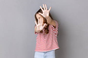 Portrait of scared little dark haired girl wearing striped T-shirt gesturing stop with palms and...