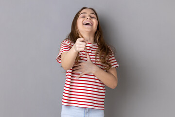 Portrait of little girl wearing striped T-shirt laughing, holding stomach and pointing to camera, taunting you, can't stop hysterical laughter. Indoor studio shot isolated on gray background.