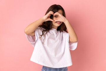 Portrait of lovely smiling little girl wearing white T-shirt showing heart shape gesture with hands and covering eye with love gesture. Indoor studio shot isolated on pink background.