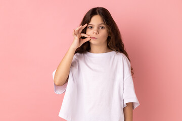 Portrait of charming little girl wearing white T-shirt with mystery look making zip gesture to close mouth, keeping secret, zipping lips. Indoor studio shot isolated on pink background. © khosrork