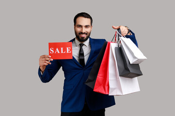 Bearded man smiling, boasting with purchases holding paper shopping bags and sale card, big holidays discounts, wearing official style suit. Indoor studio shot isolated on gray background.
